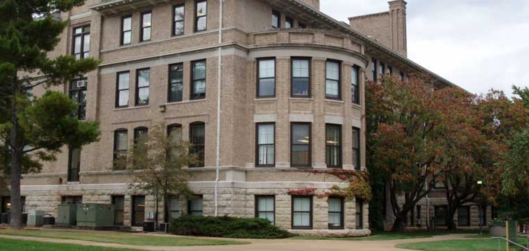 A three-story, rectangular brick building with large windows and a curved corner sits adjacent to a green, tree-lined lawn under a cloudy sky at Missouri University of Science and Technology.