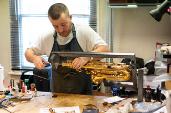 Alex Preston in the workshop above Presto's shop, repairing a saxophone held in a support on the workbench in front of him.