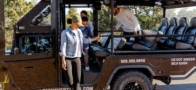 A young woman disembarks from a dark green Buck Wild Hummer Adventures tour vehicle while another person assists her.