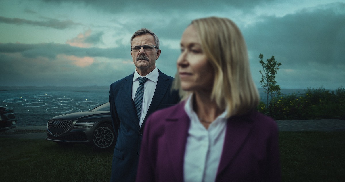 Trine Wiggen as Julie Lange stands in front of Svein Roger Karlsen as Gjert Meyer, with a gloomy sky and sea behind them in an image from the series ‘Billionaire Island.’