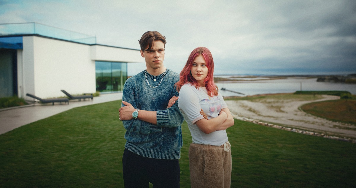 Nemi Storm as Hennie and Benjamin Bakkeid as Felix stand back-to-back on a green lawn in front of a large modern home in a still image from the series ‘Billionaire Island.’