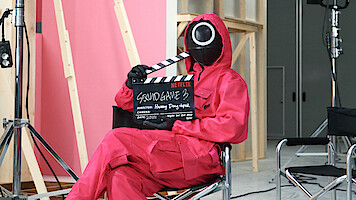 A person wearing a red jumpsuit with a black mask holds a clap board that reads "Squid Game 3"