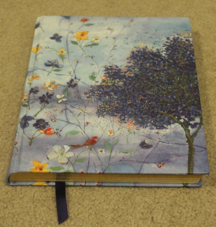 The image shows a blue hardback journal. The cover has impressionist-style art flowers, a tree, and a bay of sea.