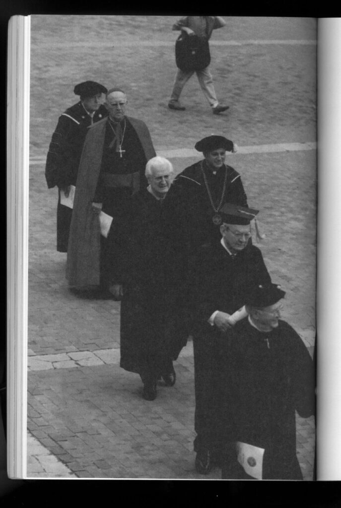 The image shows a black-and-white page from an academic catalog. It's a full page photo of six old white men in garb that is religious or academic or both: black robes, large crosses on necklaces, and so on. They are walking in a line, most of them smiling.