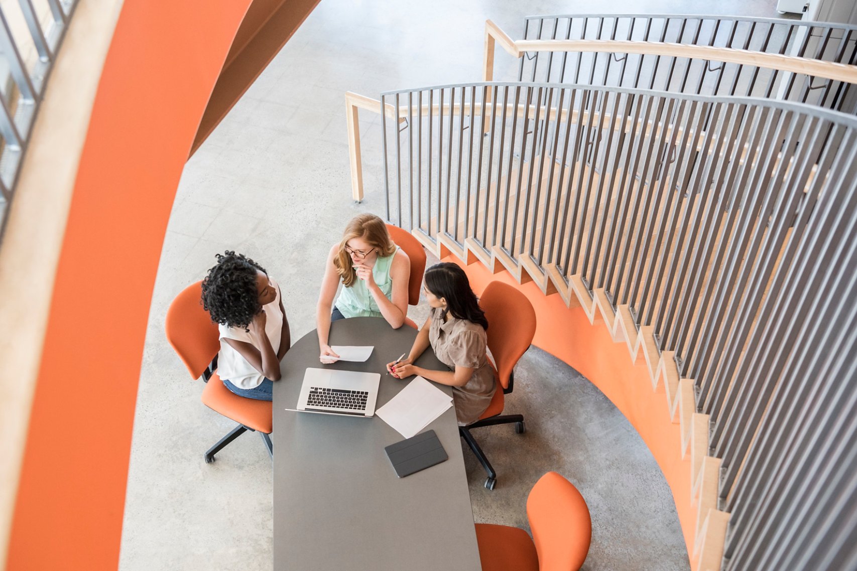 They are sitting on orange chairs and discussing together with laptop and notes