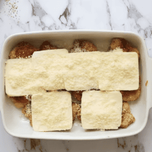 breaded fried chicken thighs topped with marinara sauce, slices of cheese, and sprinkled cheese in a baking pan.