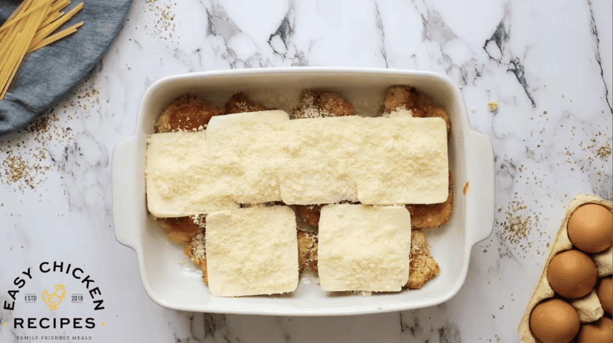 breaded fried chicken thighs topped with marinara sauce, slices of cheese, and sprinkled cheese in a baking pan.