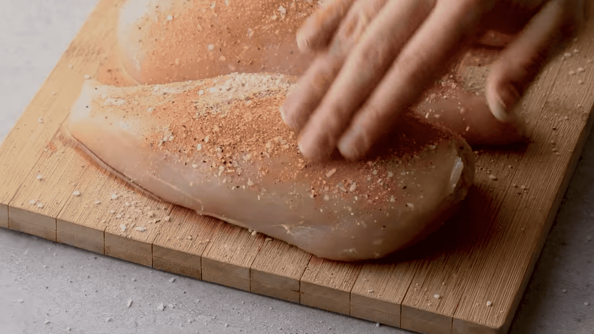 Hand rubbing spices into a raw chicken breast, placed on a cutting board.