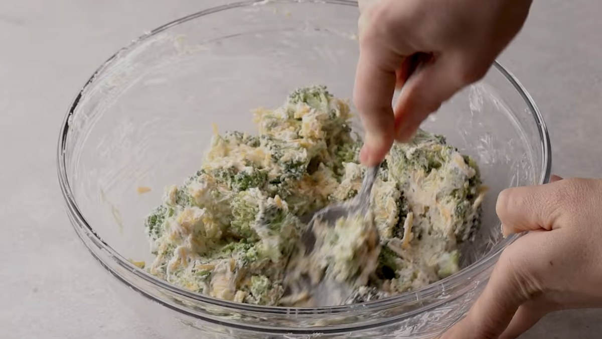 Hand mixing together broccoli, cream cheese, and shredded cheese in a glass mixing bowl.