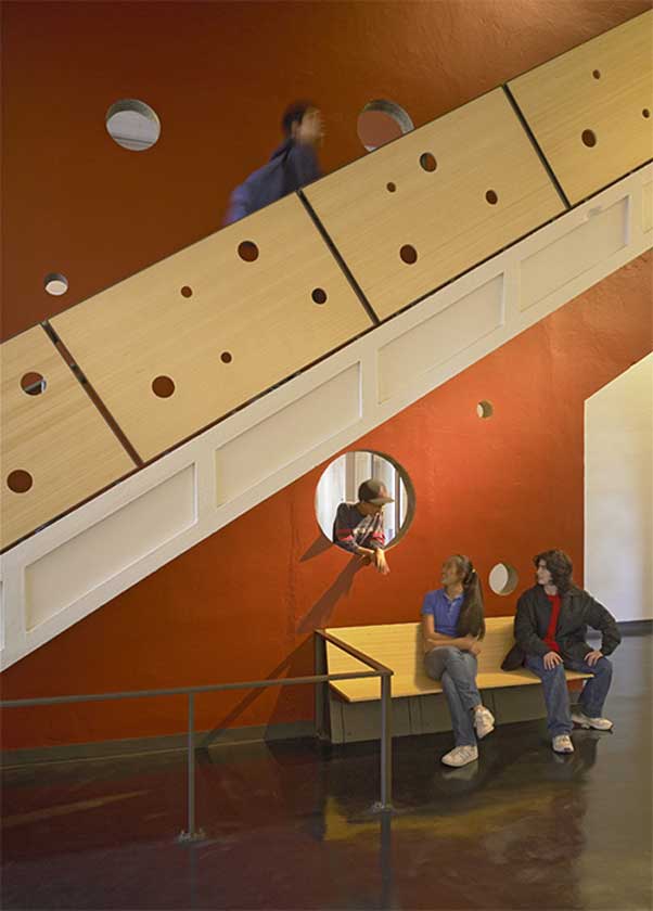 Young people chat near playful interior staircase with holes of various sizes cut in the handrail siding and walls between rooms