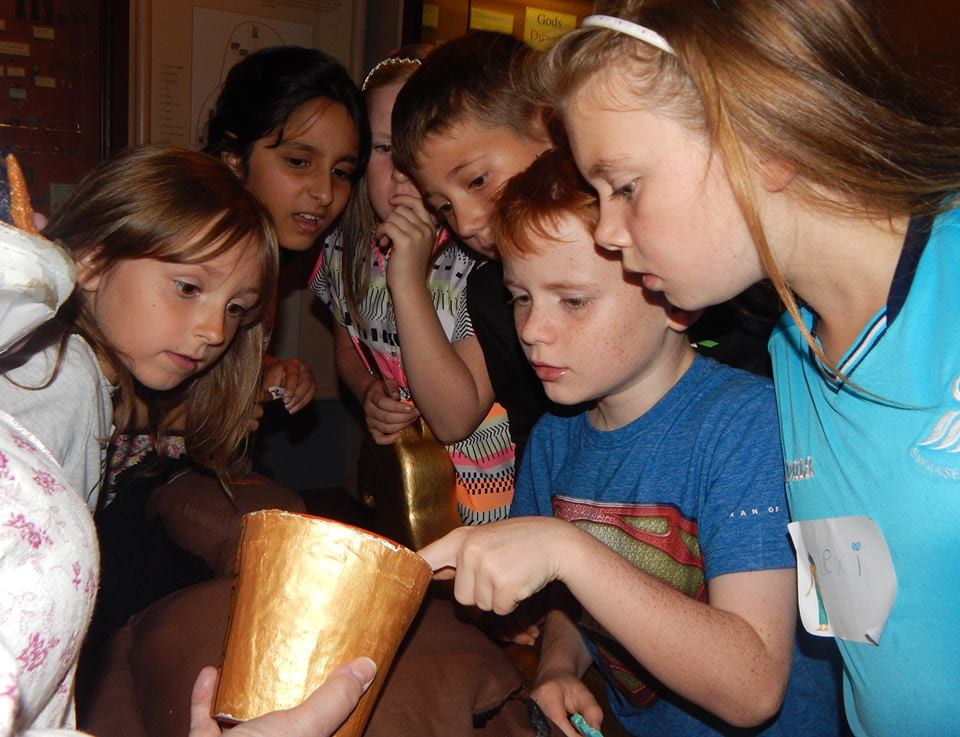 A group of children gathered closely around an object. They are looking at it with curiosity and interest, and one child is pointing at the object. 