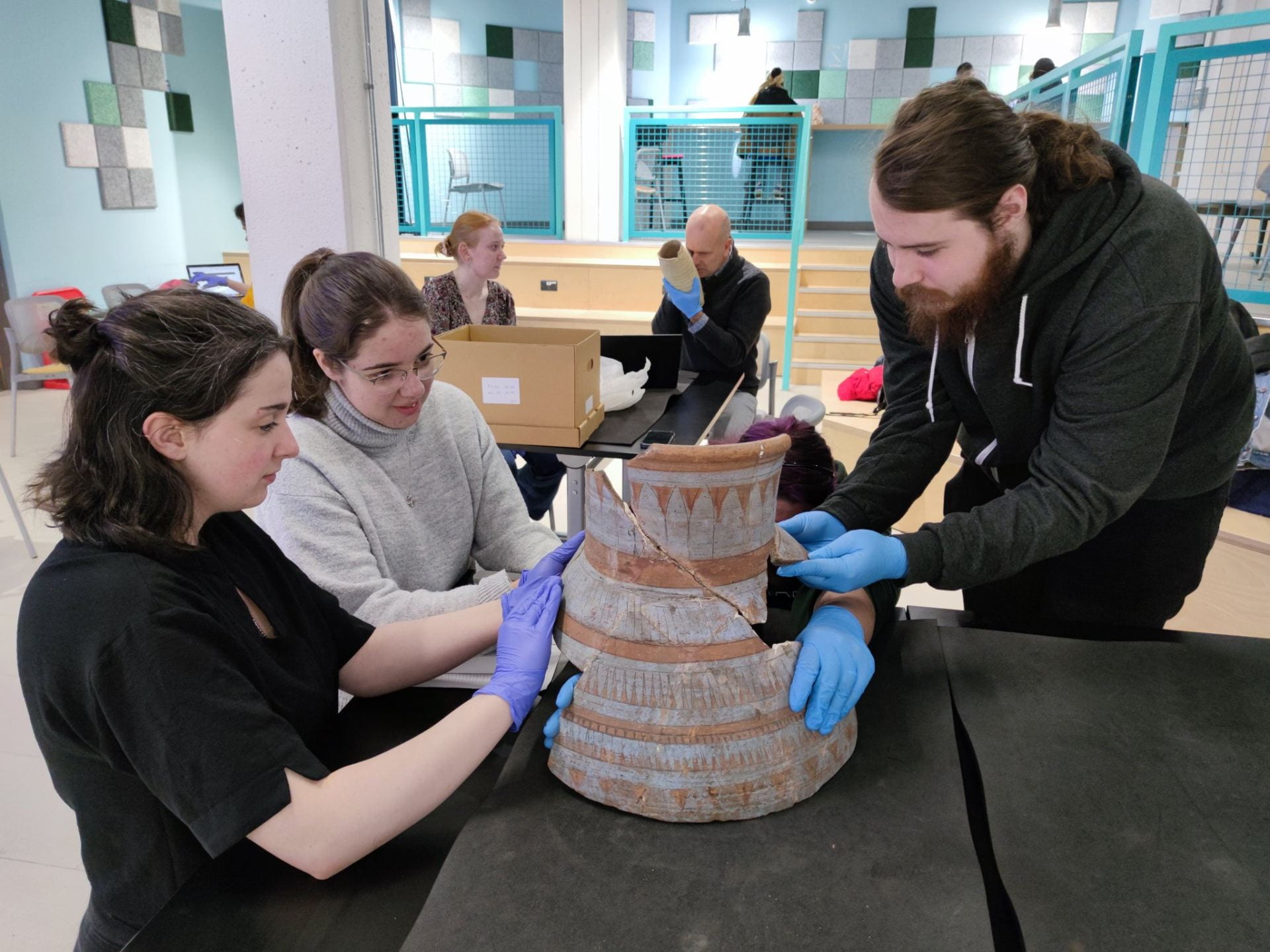 Three students, two females and one male, wearing gloves reconstructing a large pottery vessel with blue painted decoration. 