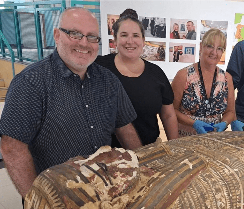 The image shows three people standing behind an ancient Egyptian coffin. From left to right, the individuals are:

Ken Griffin: A man with glasses, a short beard, and a bald head. He is smiling and wearing a dark short-sleeved shirt with a subtle pattern.
Meg Gundlach: A woman with her hair tied up in a bun, smiling warmly. She is wearing a plain black shirt.
Wendy Goodridge: A woman with glasses and blonde hair, also smiling. She is wearing a sleeveless patterned top and blue gloves, suggesting she is handling or working with the artefact.