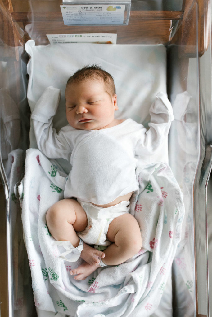 Newborn baby boy in a hospital bassinet laying on a blanket with pink and blue feet 