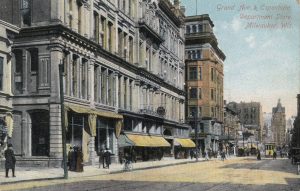 A painted postcard illustrates the Espenhain department store on the left side of Grand Avenue. The store is depicted in gray color with yellow awnings and rises among other buildings that stretch along the street. A streetcar is visible in the right background.