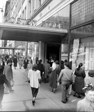 A grayscale image shows groups of people walking towards and passing the Boston Store. The shop has a sidewalk shed with store signage installed atop.