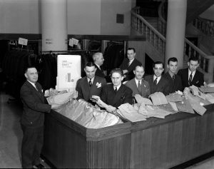 Long shot of salespeople in suits and ties standing next to tables full of collared shirts at men's shirt counter in Gimbel Bros. department store. A standing sign near the tables mentions the shirts' sale prices at $2 and $2.50.