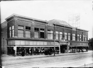 Grayscale wide shot of Lion Department store facade. The two-story rectangular building features display windows on its ground floor that showcase products such as clothes worn by mannequins. On the top center of the facade is inscribed "Goldmann." Below is the main entrance with the "Lion Store" signage atop.