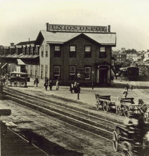 Sepia-colored elevated shot of the Union Depot on Reed Street. The building with the name sign reading Union Depot stands at the image's center-to-left background. Other buildings are visible on the far right background. Cars and horse-drawn vehicles appear next to the train tracks that stretch from the left background to the right foreground. Some people stand on the open space between the Union Depot building and the tracks.