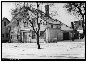 Grayscale long shot of the Lowell Damon house facing slightly to the left. The wooden frame house's facade and side are visible. The one-and-a-half-story building features two chimneys and gable roofs. The facade has an entrance in the center and a series of rectangular windows on the ground floor and above the door. A leafless tree grows in front of the house. Another one in the far right in the background. Snow covers the ground and street.