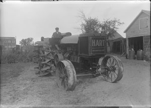 Grayscale photo of C.W. Hart on an old tractor facing slightly to the right in an outdoor space. Hart appears on the left. The vehicle's front body is inscribed "Hart, Wauwatosa. Wis." Tre tractor wheels are visible. Wooden frame buildings are on the far right.