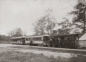 A bunch of passengers inside the Milwaukee and Whitefish Bay Dummy Line. The locomotive is on the furthest right. Three people stand on the ground next to the train and pose for this sepia-coloured photograph. Railways track lines span the surface next to the Dummy Line close to the foreground