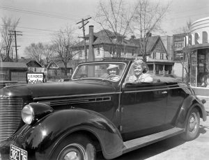 Sepia-colored wide shot of two women smiling to the camera while sitting in a car at a service station. A Wisconsin license plate is installed on the car's bottom part in the foreground. Residential and business buildings appear in the background.