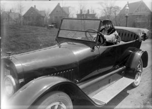 A grayscale portrait of a woman posing in the driver seat of a touring automobile that faces slightly to the left. She smiles while making eye contact with the camera lens. A small American flag adorns the windshield of the car, whose cover is down. An open grassy area is to the left of the car and large duplex houes are visible in the background.