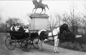 Grayscale long shot of four people sitting in a horse-drawn wagon that faces right. The passengers wear black and white clothes, and the horse is piebald. The people make direct eye contact with the camera lens. Visible behind them is the bottom part of the Kosciuszko Monument, which shows a horse statue facing left atop a tall concrete base.