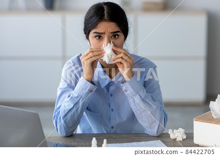 Upset sad young indian female worker suffer from allergies, colds and sickness at workplace with laptop, blowing nose 84422705