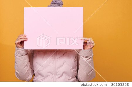 woman in pink winter jacket and hat holds blank pink sheet of paper on yellow background. Seasonal sale 86105268