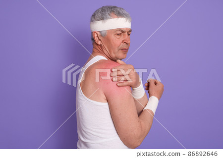 Horizontal shot of injured senior man posing sideways, mature male with white head band, elderly sportrsman posing isolated over lilac studio background, guy touching his pain shoulder, need treatment 86892646