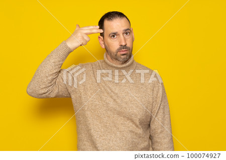 Annoyed hispanic man with beard making gun gesture with finger to head, shooting himself out of boredom or annoyance, tired of something stupid, standing over yellow background. 100074927