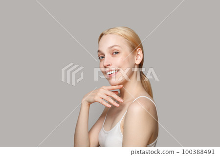 Geogeous blonde woman in white tank top with straps with ponytail smiling on grey background 100388491