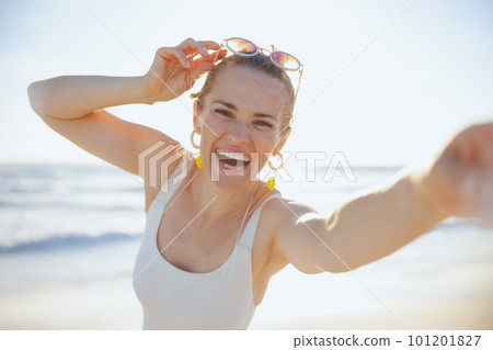 smiling stylish woman in white swimsuit at beach taking selfie 101201827