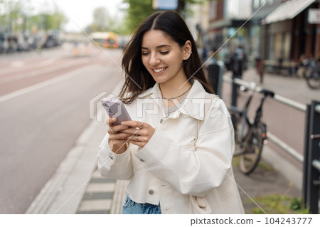 Everyday commute. Cheerful young woman student using the app on her phone while waiting for the public transport 104243777