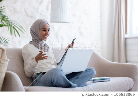 Young Muslim businesswoman in hijab working remotely, sitting on sofa at home in lotus position, holding documents in hands and smiling talking via laptop video call. 106638869