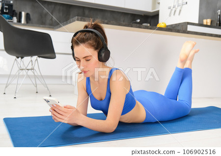 Image of young woman watches sport gym tutorials on smartphone, laying on yoga mat, doing fitness workout at home, wearing blue leggings and sportsbra 106902516