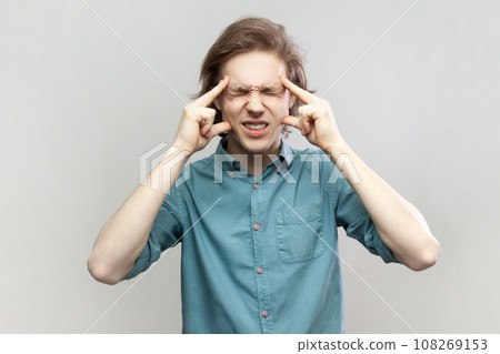 Portrait of sick ill unhealthy young man standing massaging his temples, suffering headache, frowning face, keeps eyes closed, wearing blue shirt. Indoor studio shot isolated on gray background. 108269153