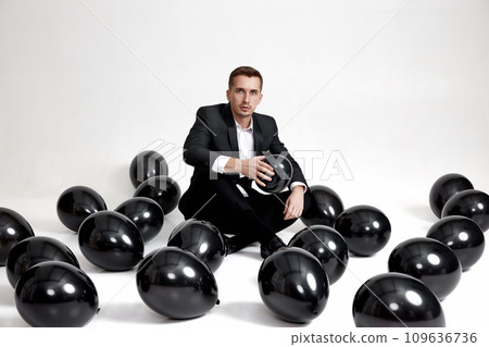 elegant caucasian man in suit tuxedo sitting on studio floor with a lot of black air balloons 109636736