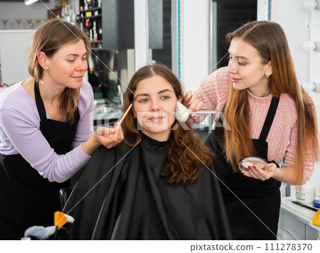 Two makeup artists apply makeup on the face of a young woman 111278370