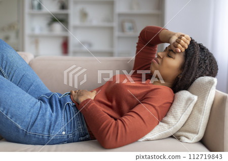 Young black woman experiencing stomach pain while lying on couch at home 112719843