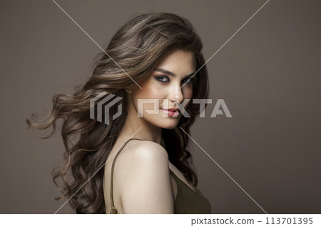 Elegant woman with long healthy wavy hair, cute smile and make-up looking at camera on brown studio 113701395