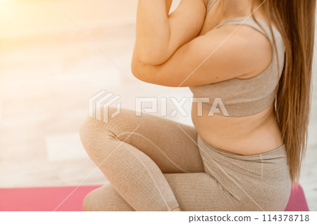 Young woman doing yoga in the gym. A girl with long hair and in a beige tracksuit stands in a cow pose on a pink carpet. A woman performs Gomukhasana. 114378718