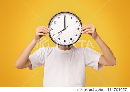 Time management concept. Teen boy covering his face with big clock, orange panorama background 114701657