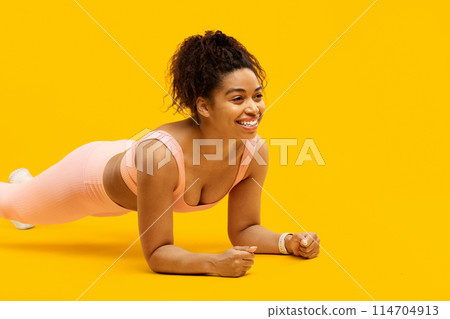 Engaged african american woman practicing a plank pose in fitness attire on an isolated yellow backdrop 114704913