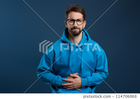 Stomach Pain. Sick young man suffering from acute abdominal ache, upset millennial guy touching belly and frowning, feeling unwell while standing against blue studio background, copy space 114706068