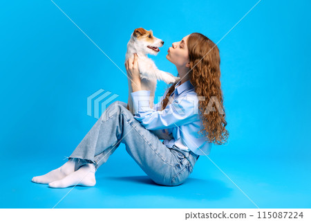 Charming, pretty girl with long hair sits on floor, kissing her lovely Jack Russel dog against vibrant blue studio background. 115087224