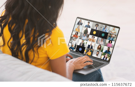 A young black woman is engaged in a virtual team meeting while working from her home office. She is seated, looking at a laptop screen displaying numerous colleagues via video call 115637672