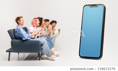 Multiracial group of young people sitting on couch, looking at huge phone with blank blue screen, isolated on white studio background, mockup, copy space 116613279